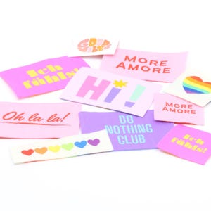 May include: A collection of rectangular fabric patches featuring various colorful designs and text including "GOOD VIBES," "Ich fühl's!," "MORE AMORE," "Hi!," "Oh la la!," "DO NOTHING CLUB," and a row of rainbow hearts and a rainbow heart shape.