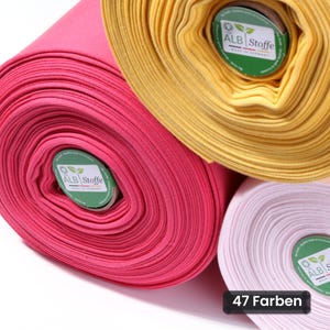 Può includere: Tre rotoli di tessuto in rosa acceso, giallo e rosa chiaro. Ogni rotolo presenta un'etichetta circolare verde con il testo ALB Stoffe Made in Germany. Il rotolo rosa chiaro include un'etichetta rettangolare nera con il testo 47 Farben.