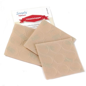 Könnte beinhalten: Drei quadratische Bögen mit transparenten, selbstklebenden runden Punkten für Lineale, beschriftet mit Linealstopper und dem Markennamen Snaply Nähkram auf der Verpackung. Jeder Bogen enthält neun kreisförmige Klebepunkte in einem Raster.