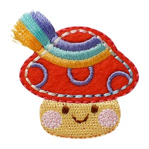 Pode incluir: Um emblema bordado de cogumelo vermelho com um rosto sorridente, bochechas rosadas, olhos castanhos e uma boca pequena. O chapéu do cogumelo é decorado com círculos roxos e um detalhe de borla arco-íris fixado no lado superior esquerdo.