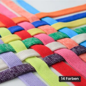 Könnte beinhalten: Ein gewebtes Gitter aus flachen Stoffstreifen in verschiedenen Farben wie Orange, Blau, Gelb, Grün, Rot, Pink, Weiß, Violett und hellem Lavendel. Die Streifen sind eng miteinander verflochten. Ein schwarzes rechteckiges Etikett zeigt den Text 14 Farben.