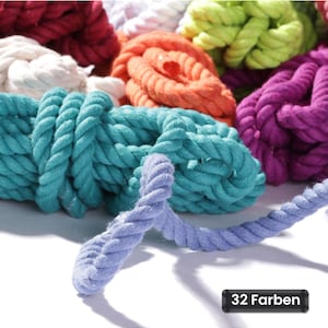 Peut inclure: Une collection de cordes en coton torsadé de diverses couleurs vives, notamment turquoise, violet, orange, vert lime, blanc et rouge. Un brin de corde bleu clair s'étend vers l'avant. Une étiquette noire rectangulaire affiche le texte 32 Farben.