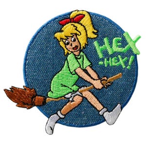 Pode incluir: Um emblema bordado circular apresentando uma personagem de desenho animado com cabelo loiro e um laço vermelho, vestindo uma camisa verde, montada em uma vassoura marrom. O fundo é azul e o texto HEX-HEX! é bordado em fio verde brilhante.