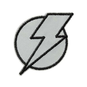 Könnte beinhalten: Ein kreisförmiger gestickter Aufnäher mit hellgrauer Mitte und einem dicken schwarzen Rand. Ein gezacktes Blitzsymbol ist in das Design integriert, mit schwarzem Garn umrandet und erstreckt sich über die kreisförmige Basis bis zum Rand.
