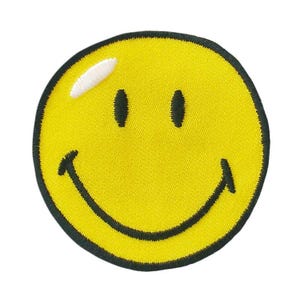 Pode incluir: Um emblema bordado circular com uma cara sorridente amarela brilhante e uma borda preta. O design inclui dois olhos ovais pretos, uma boca larga e curva preta e uma pequena marca branca no lado superior esquerdo do rosto.
