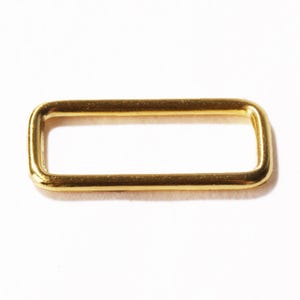 Op de afbeelding: Een rechthoekige metalen lus met een gladde, gepolijste goudkleurige afwerking. Het beslag heeft afgeronde hoeken en een hol midden, ontworpen als een functionele verbinding voor riemen of banden in diverse knutselprojecten.