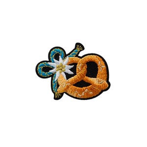 Könnte beinhalten: Ein gestickter Aufnäher mit einer goldbraunen Brezel, die mit einer weißen Edelweißblüte und einer blauen geflochtenen Kordel verschlungen ist. Das Motiv ist mit schwarzem Garn umrandet und weist detaillierte Stickereien auf.