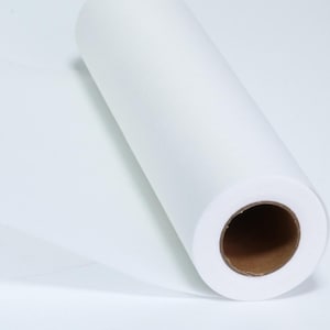Op de afbeelding: Een grote rol wit papier of stofmateriaal dat strak om een cilindrische bruine kartonnen kern is gewikkeld. Het materiaal is ondoorzichtig, glad en heeft een matte textuur, en steekt iets uit de rol om een plat, dun veloppervlak te tonen.