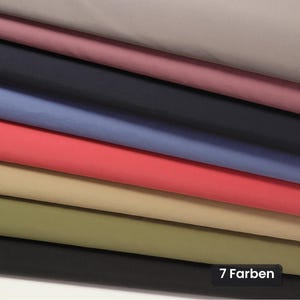 Pode incluir: Uma pilha de sete peças de tecido dobradas em várias cores sólidas, incluindo cinza, rosa empoeirado, azul marinho escuro, azul claro, vermelho coral, bege e verde oliva. Uma pequena etiqueta retangular preta no canto inferior direito exibe o texto 7 Farben.