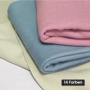 Op de afbeelding: Drie gevouwen lagen stof in pasteltinten van lichtgeel, stoffig blauw en gedempt roze. De stof heeft een matte, viltachtige textuur. Een klein zwart rechthoekig label in de rechterbenedenhoek toont de tekst 14 Farben.