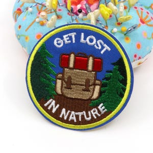 Pode incluir: Um emblema bordado circular com fundo azul e um gráfico central de uma mochila bege e vermelha. O texto GET LOST IN NATURE está bordado em letras brancas. Pinheiros verde-escuros ladeiam a mochila sobre um solo castanho com borda amarela.