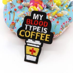 Może przedstawiać: Czarna haftowana naszywka w kształcie kubka do kawy z napisem MY BLOOD TYPE IS COFFEE wykonanym czerwoną, białą i żółtą nicią. Kubek posiada żółtą opaskę z symbolem czerwonego krzyża, a naszywka spoczywa na jasnoniebieskiej poduszce na szpilki.