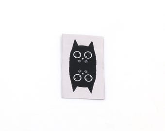 May include: A rectangular woven fabric label featuring a light grey background with a black graphic of two cat heads mirrored vertically. Each cat head has large white circular eyes, a small white nose, and thin white whiskers on a black silhouette.