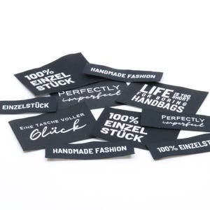 Könnte beinhalten: Eine Ansammlung schwarzer Stoffetiketten mit weißem Text, darunter Phrasen wie 100% EINZELSTÜCK, HANDMADE FASHION, PERFECTLY imperfect, LIFE IS TOO SHORT FOR BORING HANDBAGS und EINE TASCHE VOLLER Glück, die in einem Haufen angeordnet sind.