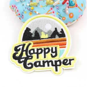 Pode incluir: Um emblema bordado com uma paisagem de montanha, uma tenda amarela, um sol e faixas horizontais em laranja e castanho. O texto Happy Camper está bordado em letras cursivas pretas na parte inferior do design circular.