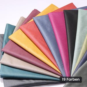 Op de afbeelding: Een waaiervormige opstelling van stofstalen in diverse kleuren, waaronder geel, rood, blauw, roze, zwart, grijs en groen. De collectie heeft een gladde, matte textuur en bevat een zwart label met de tekst 19 Farben.