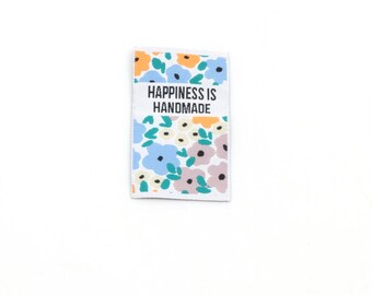 May include: A rectangular fabric clothing label featuring a white background with a repeating floral pattern in shades of light blue, orange, and muted purple, with green leaves. The text "HAPPINESS IS HANDMADE" is printed in black.