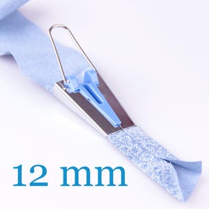 Könnte beinhalten: Ein 12 mm Schrägbandformer mit einem silberfarbenen Metallgehäuse, einem hellblauen Kunststoffeinsatz und einem Drahtgriff. Das Werkzeug ist abgebildet, wie es einen hellblauen Stoffstreifen faltet, um ein Schrägband zu formen.