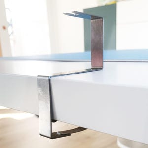 Könnte beinhalten: Eine silberfarbene Metallklemme mit flachem, L-förmigem Design und einer eingekerbten Oberkante. Die Halterung besteht aus einem einzigen Stück gebogenem Metall und ist so konzipiert, dass sie zur Montage oder Unterstützung über die Kante einer Fläche greift.