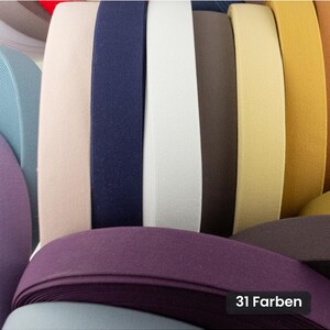 Op de afbeelding: Een collectie stoffen elastische banden gerangschikt in een verticale rij, met een verscheidenheid aan kleuren waaronder lichtroze, marineblauw, wit, bruin, geel en paars. Een zwart rechthoekig label in de hoek toont de tekst 31 Farben.