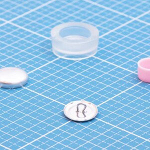 Könnte beinhalten: Ein Set aus Bastelkomponenten, bestehend aus einer transparenten Silikonform, einem kleinen silberfarbenen Metallknopf-Rückteil mit Öse, einer glatten silberfarbenen Knopfkappe und einem kleinen rosa zylindrischen Behälter auf einer blauen Rastermatte.