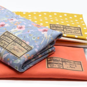 Könnte beinhalten: Ein Stapel gefalteter Stoffe, bestehend aus einem blauen Softshell-Stoff mit rosa Blumenmuster, einem gelben Baumwollstoff mit weißen Punkten und einem einfarbigen orangefarbenen Baumwollstoff. Jedes Stück hat ein rechteckiges Papieretikett.