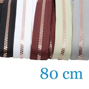 Könnte beinhalten: Eine Auswahl von fünf 80 cm langen Reißverschlüssen mit metallischen roségoldenen Zähnen und Schiebern. Die Stoffbänder sind in den Farben Schwarz, Weiß, Dunkelrot, Creme und Hellgrau gehalten und nebeneinander angeordnet, um die Auswahl zu zeigen.