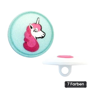 Può includere: Un bottone rotondo con sfondo azzurro e una grafica di un unicorno rosa. Il bottone presenta un occhiello bianco sul retro per l'attacco. Una piccola etichetta rettangolare nera con testo bianco riporta 7 Farben, indicando sette colori.