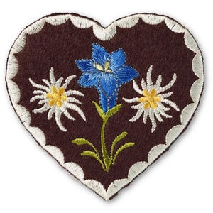 Può includere: Una toppa in tessuto marrone scuro a forma di cuore con un bordo smerlato ricamato in bianco. Il centro presenta un fiore blu ricamato a forma di campana con steli e foglie verdi, affiancato da due fiori di stella alpina bianchi con centri gialli.