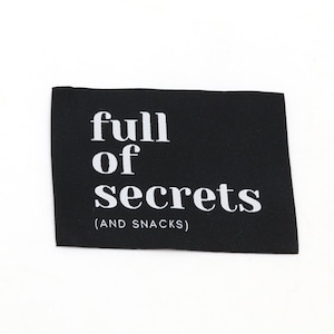 Puede incluir: Una etiqueta rectangular de tela negra con texto blanco que dice full of secrets (AND SNACKS) en una fuente serif negrita. La etiqueta tiene una apariencia plana y limpia con bordes definidos y un diseño de alto contraste para prendas.