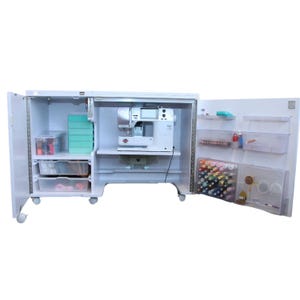 Könnte beinhalten: Ein weißer Nähschrank mit geöffneten Türen, ausgestattet mit einer zentralen Nähmaschine, mehreren transparenten Schubladen auf der linken Seite sowie verschiedenen Ablagen und Fächern an der rechten Tür für Garnrollen und Nähzubehör.