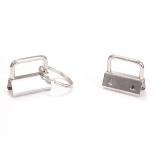 Può includere: Due componenti in metallo color argento per portachiavi, ciascuno dotato di un morsetto rettangolare con un occhiello superiore piatto. Un pezzo ha un anello circolare diviso attaccato all'occhiello, mentre l'altro è mostrato senza anello.