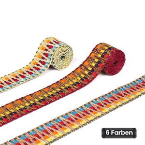 Op de afbeelding: Drie rollen decoratief geweven lint met ingewikkelde geometrische patronen in levendige veelkleurige draden. De linten tonen een herhalend vierkant en rechthoekig motief in tinten rood, geel, blauw en groen. Een zwart label bevat de tekst 6 Farben.