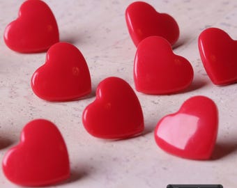 May include: A collection of small, glossy, bright red heart-shaped buttons arranged in a scattered pattern. Each button features a smooth, rounded surface and a vibrant red finish. A small black rectangular label in the corner displays the text 7 Farben.