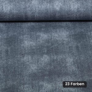 Op de afbeelding: Een rol donkerblauwe stof met een gevlekte, verweerde textuur en subtiele kleurvariaties. Het oppervlak heeft een zachte, doorleefde uitstraling met een zichtbaar weefpatroon. Een klein zwart rechthoekig label toont de tekst 23 Farben.