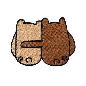 Könnte beinhalten: Ein gestickter Aufnäher mit zwei stilisierten Bären, die sich umarmen. Ein Bär ist hellbeige und der andere mittelbraun. Beide Bären haben kleine abgerundete Ohren, einfache schwarze gestickte Gesichtszüge und ein kleines schwarzes Pfoten-Detail.