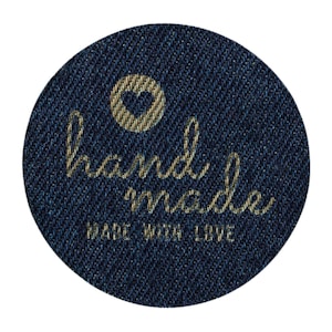Pode incluir: Um emblema circular de tecido jeans azul escuro com um ícone de coração bordado em dourado acima do texto em cursivo "hand made" e o texto em letras maiúsculas "MADE WITH LOVE" impresso em cor dourada na parte inferior do emblema.