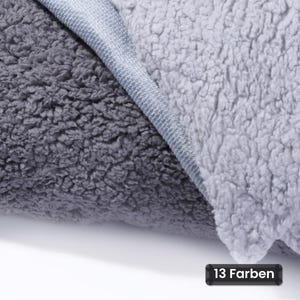 Könnte beinhalten: Eine zweifarbige Decke mit einer dunkel anthrazitfarbenen, strukturierten Fleece-Seite und einer helleren silbergrauen Fleece-Seite, getrennt durch eine Naht. Ein kleines schwarzes rechteckiges Etikett zeigt den Text 13 Farben.