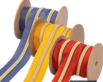 Könnte beinhalten: Drei Spulen mit Endlosreißverschluss in Blau, Gelb und Rot, jeweils mit einer metallisch goldfarbenen Reißverschlusskette mittig auf dem Stoff. Die Spulen bestehen aus brauner Pappe. Ein schwarzes rechteckiges Etikett zeigt den Text 36 Farben.