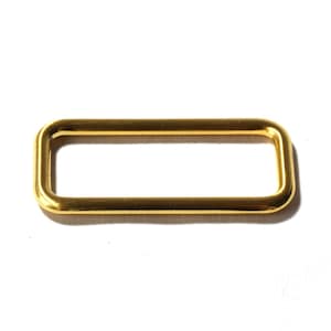 Könnte beinhalten: Eine rechteckige Metallschlaufe mit polierter goldfarbener Oberfläche. Das Beschlagteil hat abgerundete Ecken und eine glatte, durchgehende Oberfläche, die als funktionales Element für Taschen, Riemen oder Gürtel konzipiert ist.