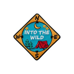 Pode incluir: Um emblema bordado em forma de diamante com uma borda preta. O centro apresenta um círculo azul com uma tenda vermelha, pinheiros verdes, uma lua crescente amarela e estrelas. O texto INTO THE WILD está em branco, com N, S, E, W em preto.