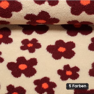 May include: A cream-colored sherpa fleece fabric featuring a repeating pattern of dark maroon flowers with bright orange centers. The fabric has a thick, textured pile and includes a small black rectangular label with the text 5 Farben.