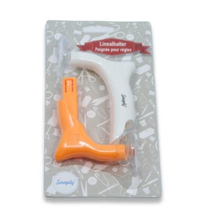 Op de afbeelding: Een wit en oranje plastic liniaalhouder verpakt op een grijze kaart met naaipictogrammen. Het product is gelabeld als Linealhalter Poignée pour règles en bevat de merknaam Snaply op het handvat en onderaan de verpakkingskaart.