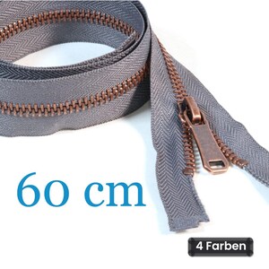 Könnte beinhalten: Ein grauer Reißverschluss mit kupferfarbenen Zähnen und einem passenden Metallschieber. Das Reißverschlussband hat ein Fischgrätenmuster. Der Text 60 cm ist in Blau dargestellt, und ein schwarzes Etikett zeigt 4 Farben in Weiß.