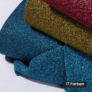 May include: Three folded layers of glitter fabric in shades of deep blue, gold, and dark red. The material has a highly reflective, textured surface with fine sparkling particles throughout. A small black rectangular label displays the text 17 Farben.