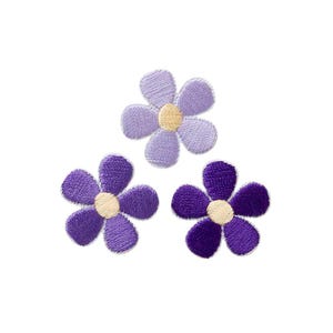Könnte beinhalten: Drei gestickte Blumenaufnäher mit jeweils fünf abgerundeten Blütenblättern. Der obere Aufnäher ist helllavendel, der untere linke ist mittelviolett und der untere rechte ist dunkelviolett. Jede Blume hat eine kreisförmige hellgelbe Mitte.
