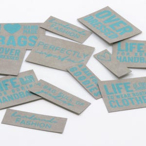 Può includere: Una collezione di etichette rettangolari in carta grigia con testo e grafiche stampate in blu brillante. Le frasi includono "Handmade Fashion", "Over Dressed", "Perfectly Imperfect", "Life is too short for boring handbags" e "A bag full of goodies".