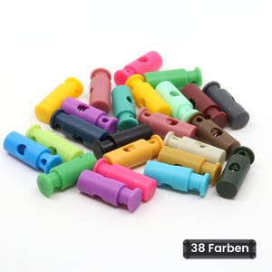 Pode incluir: Uma coleção de pequenos fechos de cordão cilíndricos de plástico em várias cores sólidas, incluindo azul, verde, amarelo, rosa, roxo, preto, castanho e bege. Cada fecho possui um mecanismo de mola com um orifício central.