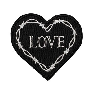 Könnte beinhalten: Ein herzförmiger gestickter Aufnäher mit schwarzem Hintergrund und weißem Rand. In der Mitte befindet sich das Wort LOVE in weißen Großbuchstaben, umgeben von einem weißen, gestickten Stacheldrahtmuster, das der Herzform folgt.