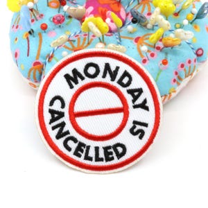 Pode incluir: Um emblema bordado circular com fundo branco e borda vermelha. O texto MONDAY IS CANCELLED está costurado em letras pretas ao redor de uma linha horizontal vermelha dentro de um círculo vermelho. O emblema repousa sobre uma almofada de alfinetes floral azul-clara.